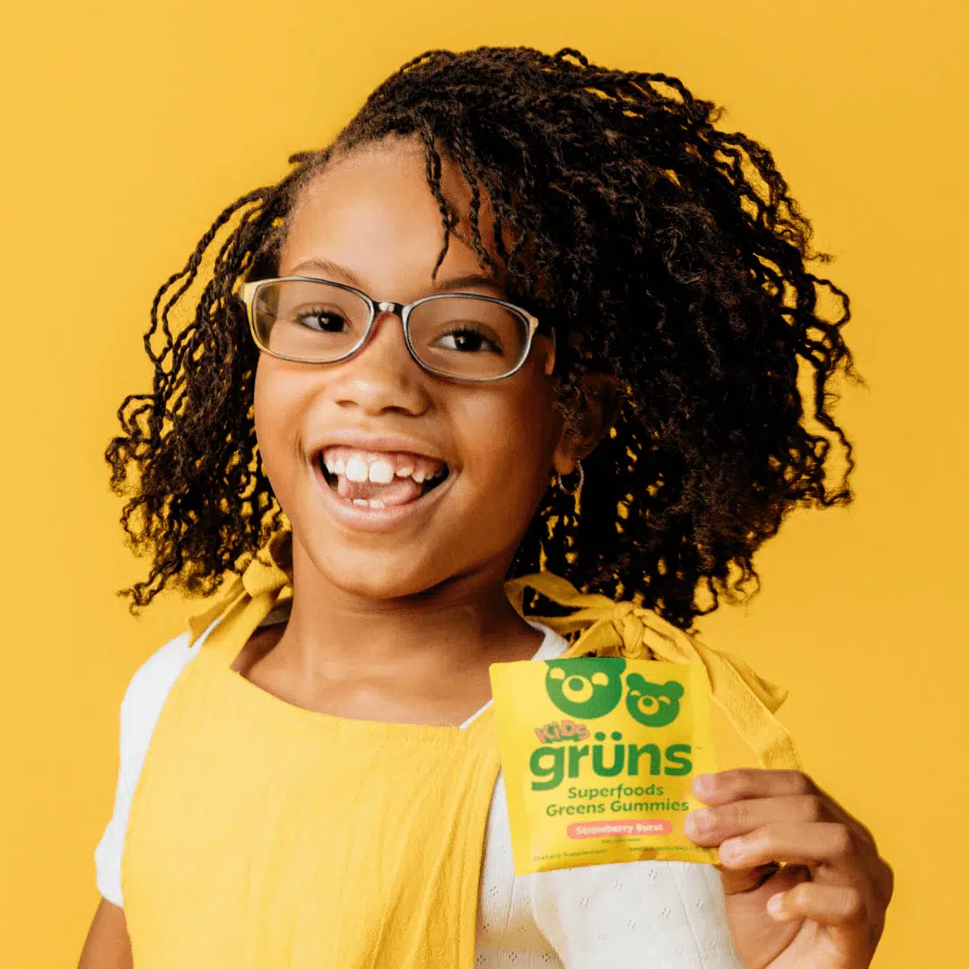 Smiling child with glasses and braided hair holds a packet of Grüns Superfoods Greens Gummies against a yellow background.