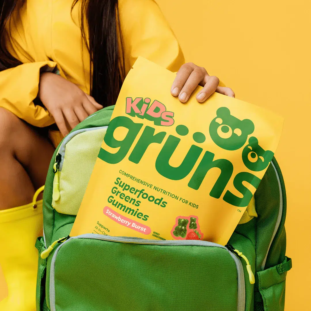 A person places a yellow bag of Kids Grüns Superfoods Greens Gummies into a green backpack against a yellow background.
