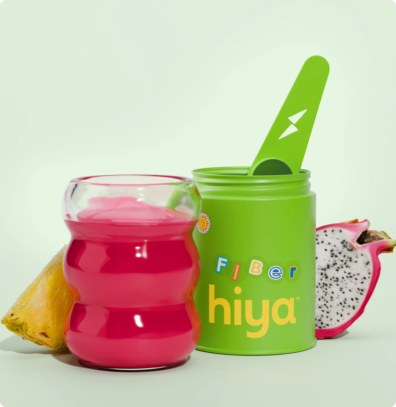 A glass of bright pink juice sits next to a green container labeled "hiya fiber" with a green spoon, plus a slice of pineapple and half a dragon fruit.