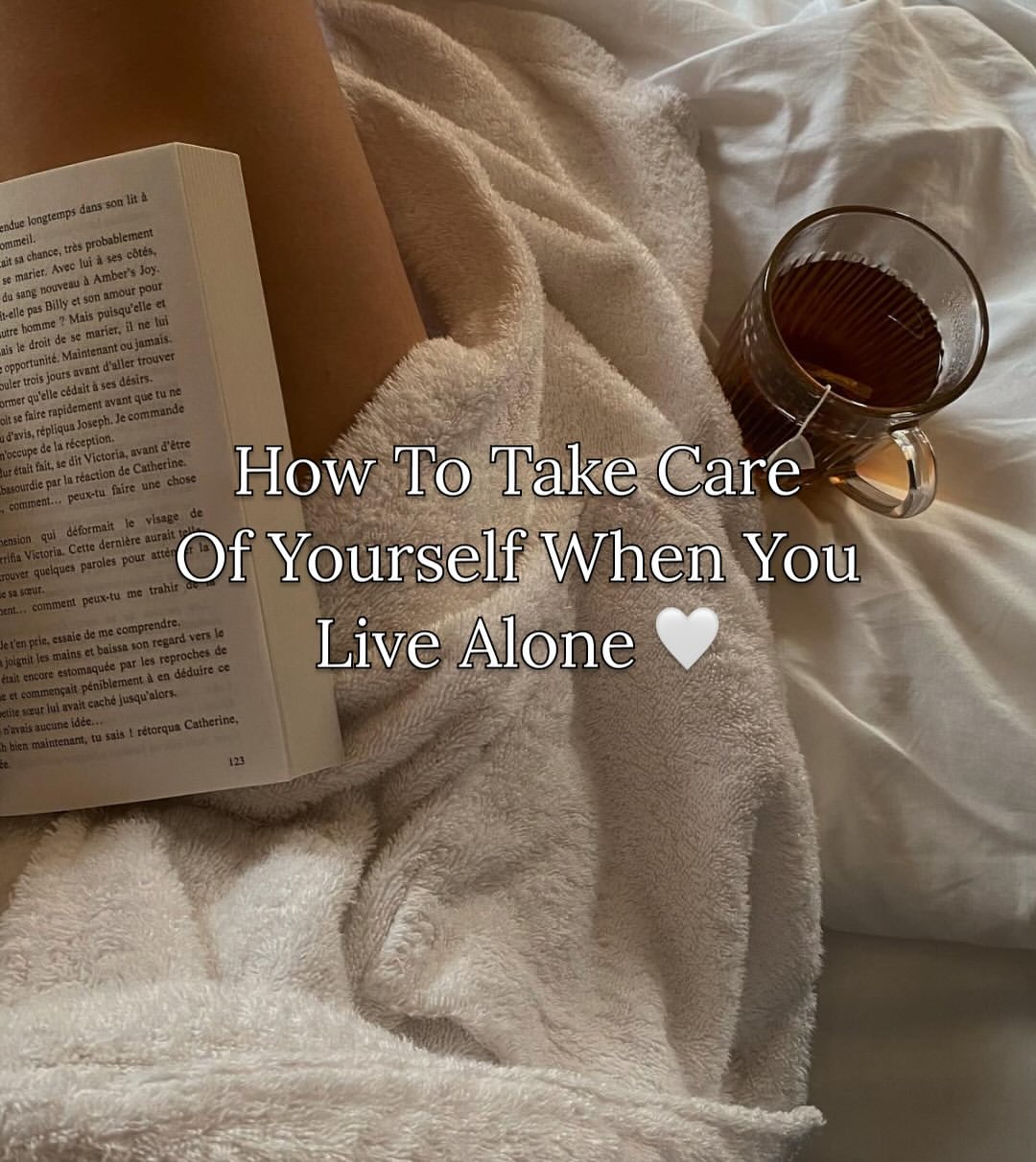 A person covered in a beige blanket sits with a book and a glass cup of tea on a bed. Text reads, "How To Take Care Of Yourself When You Live Alone.