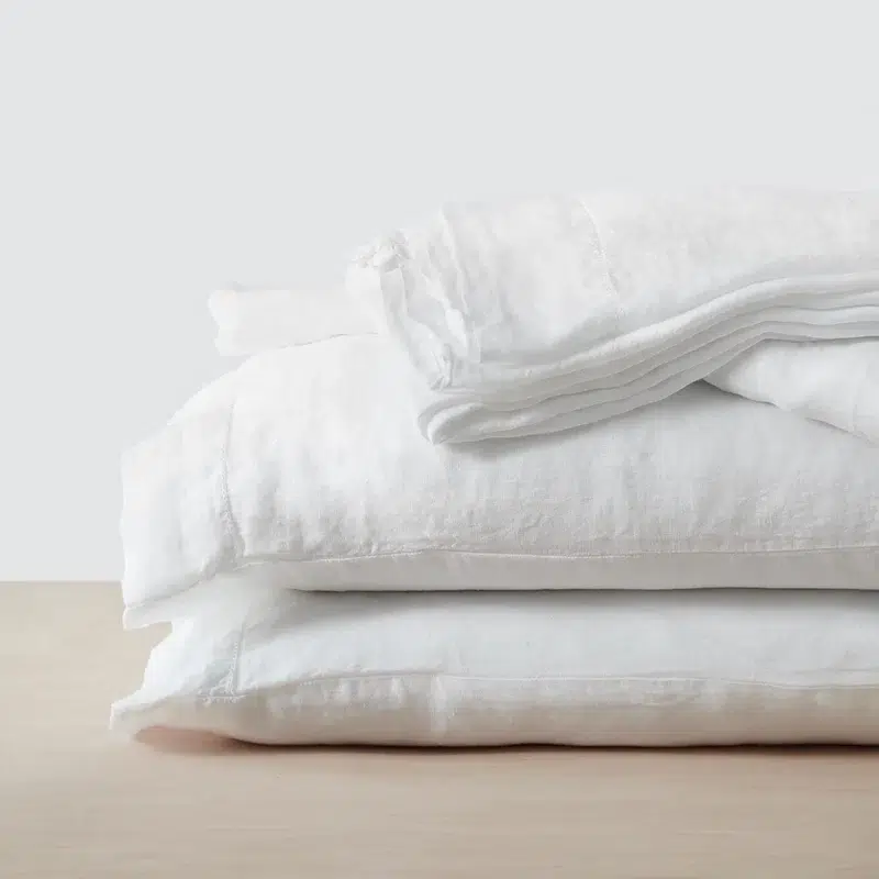 A neatly folded stack of white linen bedding, including sheets and pillowcases, placed on a light wooden surface against a plain background.
