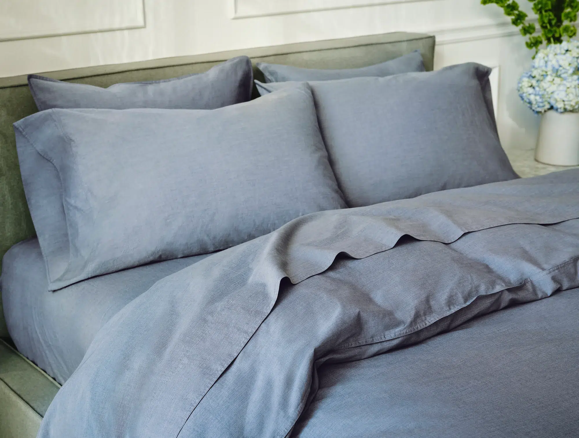 A neatly made bed with light blue-gray sheets, pillowcases, and a duvet cover, set against a light-colored headboard and white paneled wall.