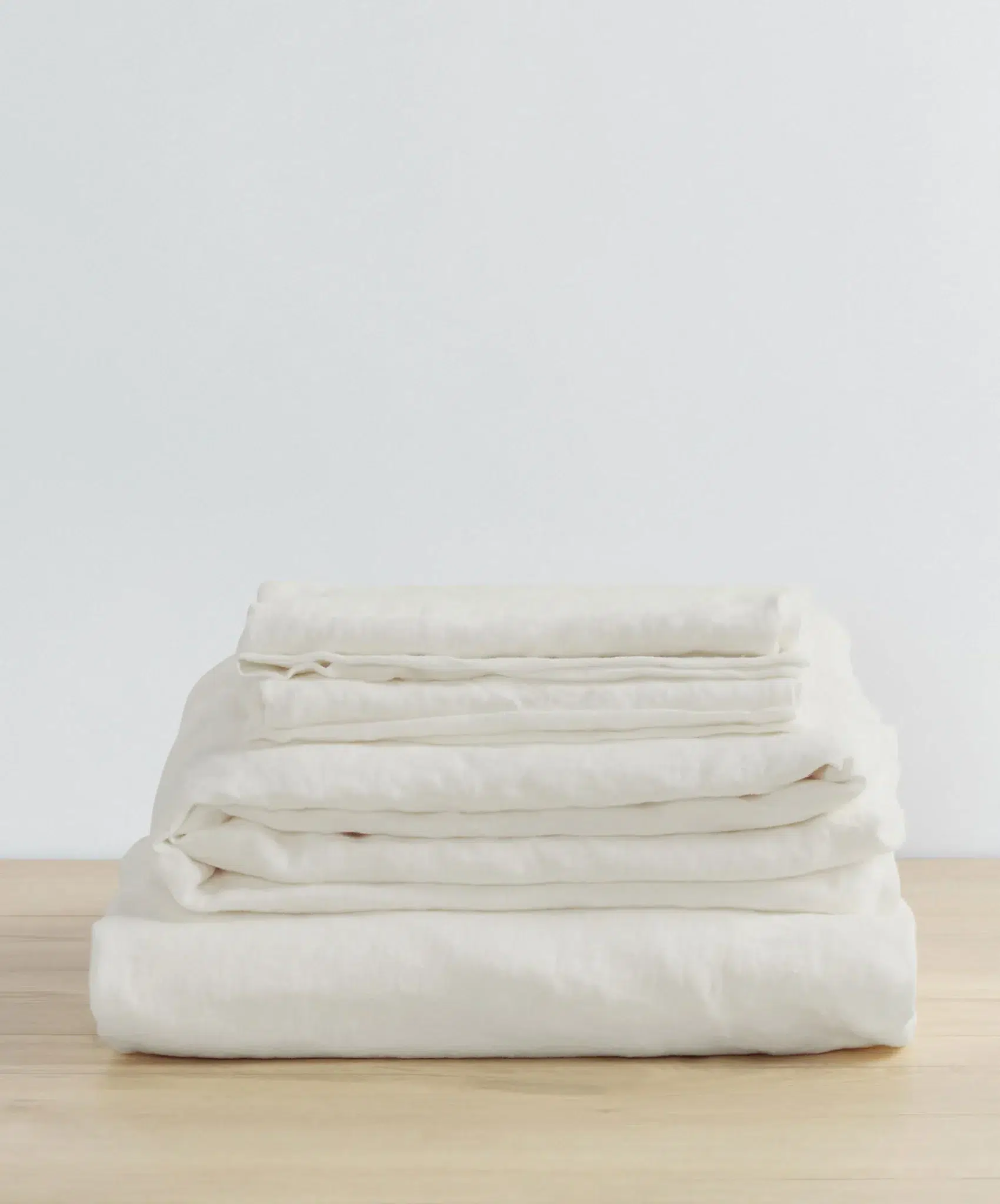 A neatly folded stack of white bed sheets sits on a light wooden surface against a plain white background.