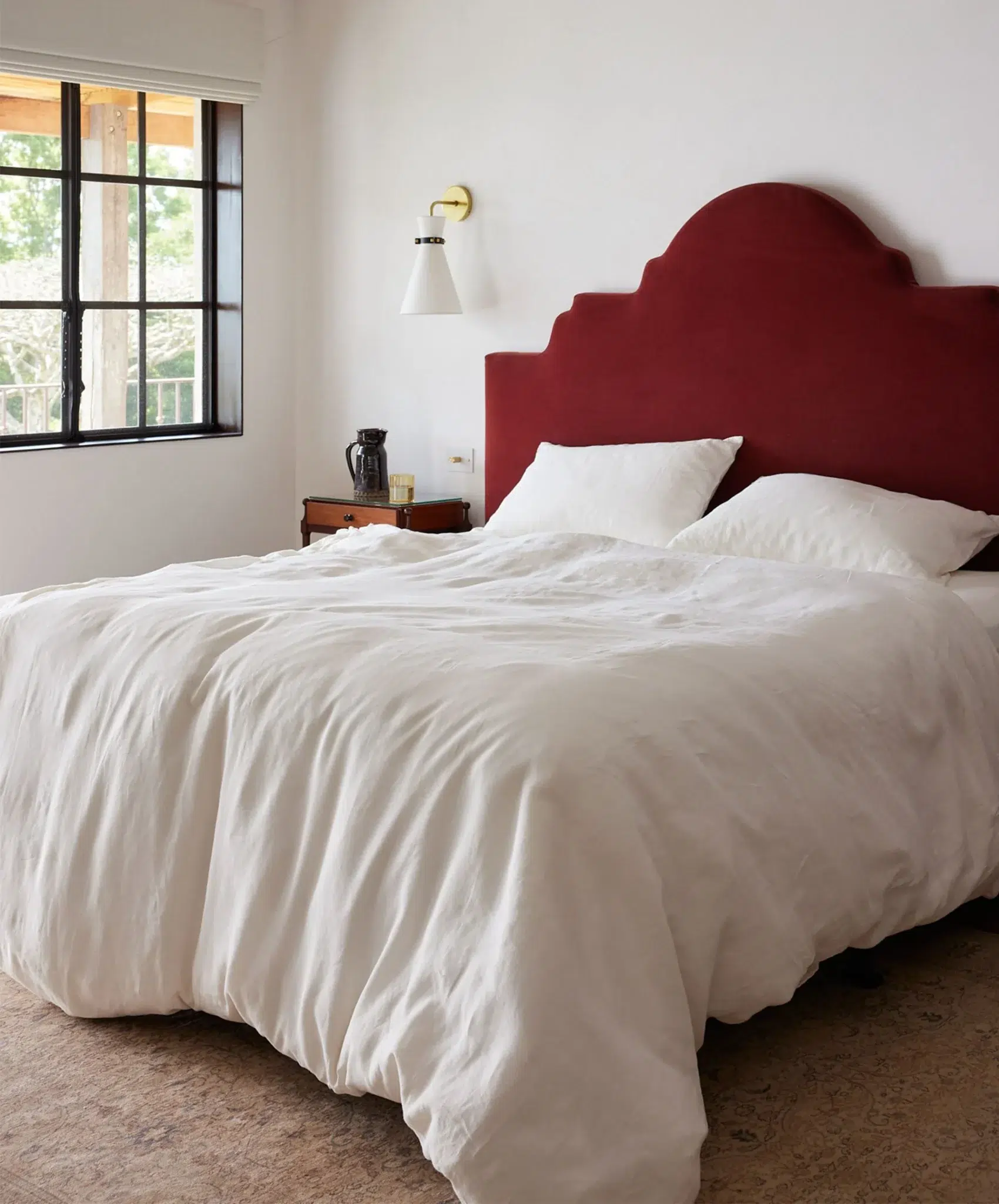 A neatly made bed with white bedding, two pillows, and a red upholstered headboard is beside a wooden nightstand and a window with black frames.