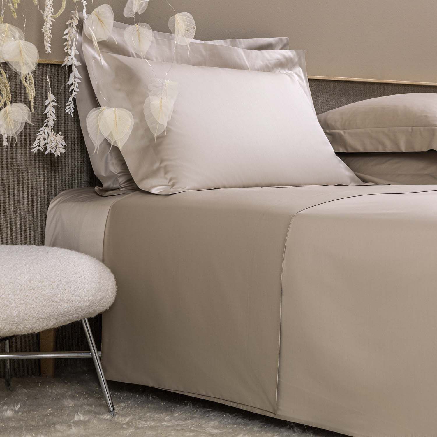 A neatly made bed with light beige sheets and pillowcases, next to a round upholstered stool and decorative white hanging leaves.