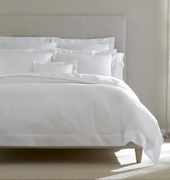 A neatly made bed with white linens and multiple pillows, set against a light gray upholstered headboard in a bright, neutral-colored room.