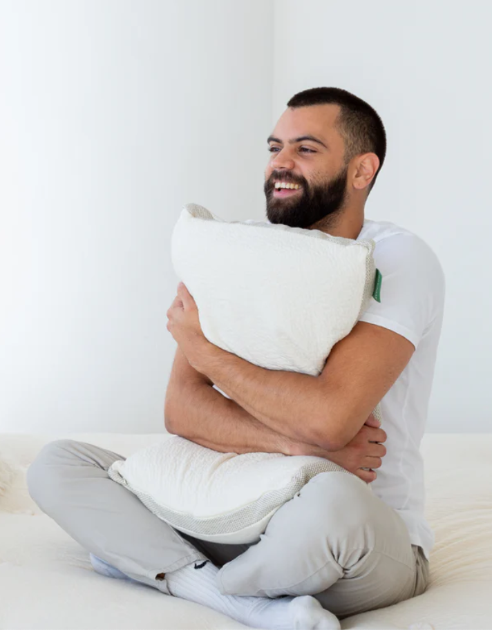 A bearded man sits cross-legged on a bed, smiling and hugging a white pillow in a brightly lit room.