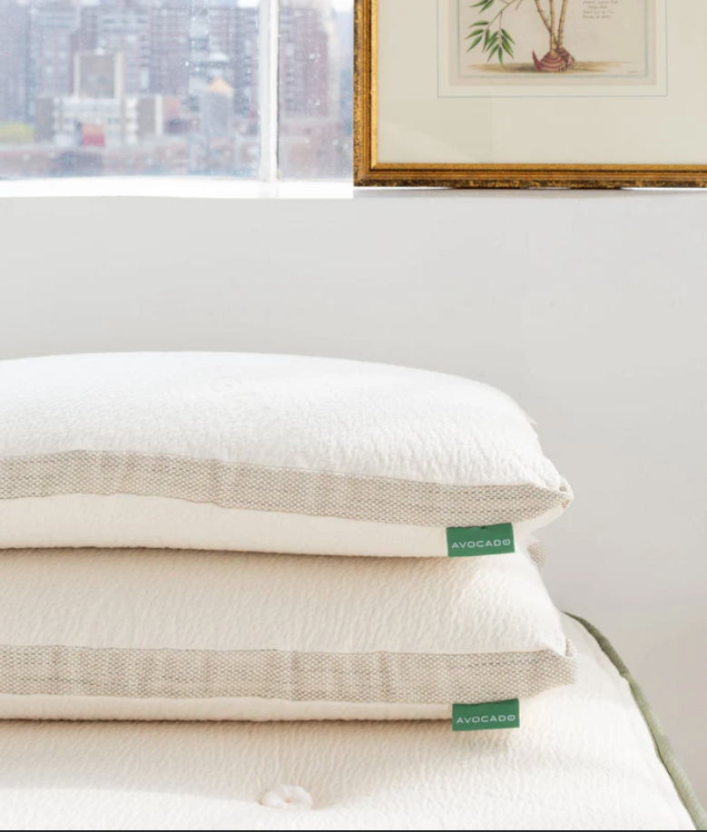 Two white pillows with green "Avocado" tags are stacked on a matching mattress near a window and a framed botanical print.