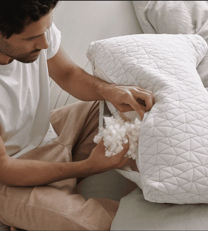 A person adjusts the filling inside a white, zippered pillow while sitting on a bed.
