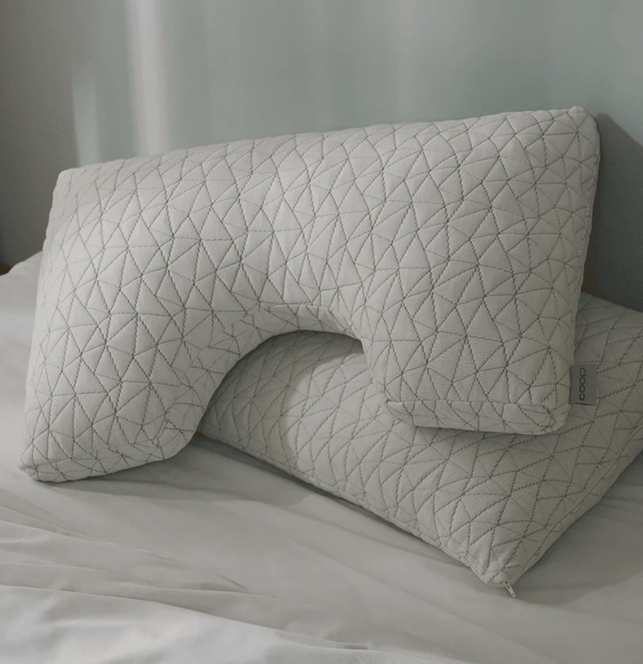 A white, U-shaped pillow with a geometric pattern is stacked on top of a standard rectangular pillow on a bed with white sheets.