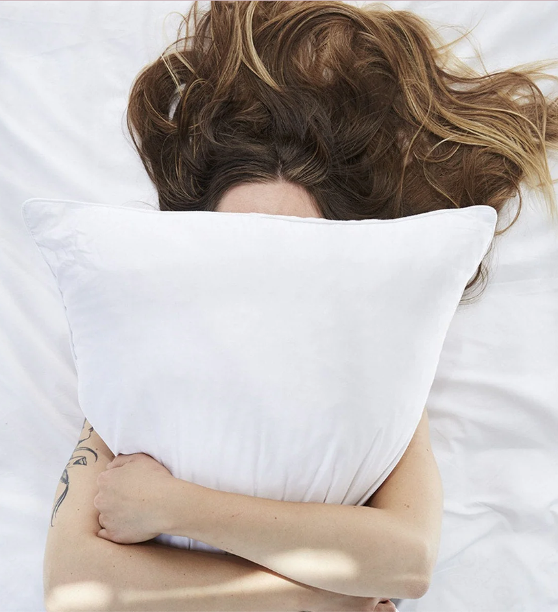 Person lying on a bed with tousled hair, hugging a white pillow and partially covering their face.