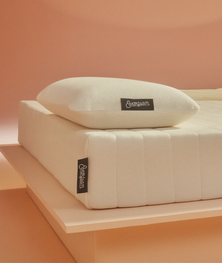 A white mattress and pillow with black "Earthfoam" labels are displayed on a light wooden platform against a peach-colored background.