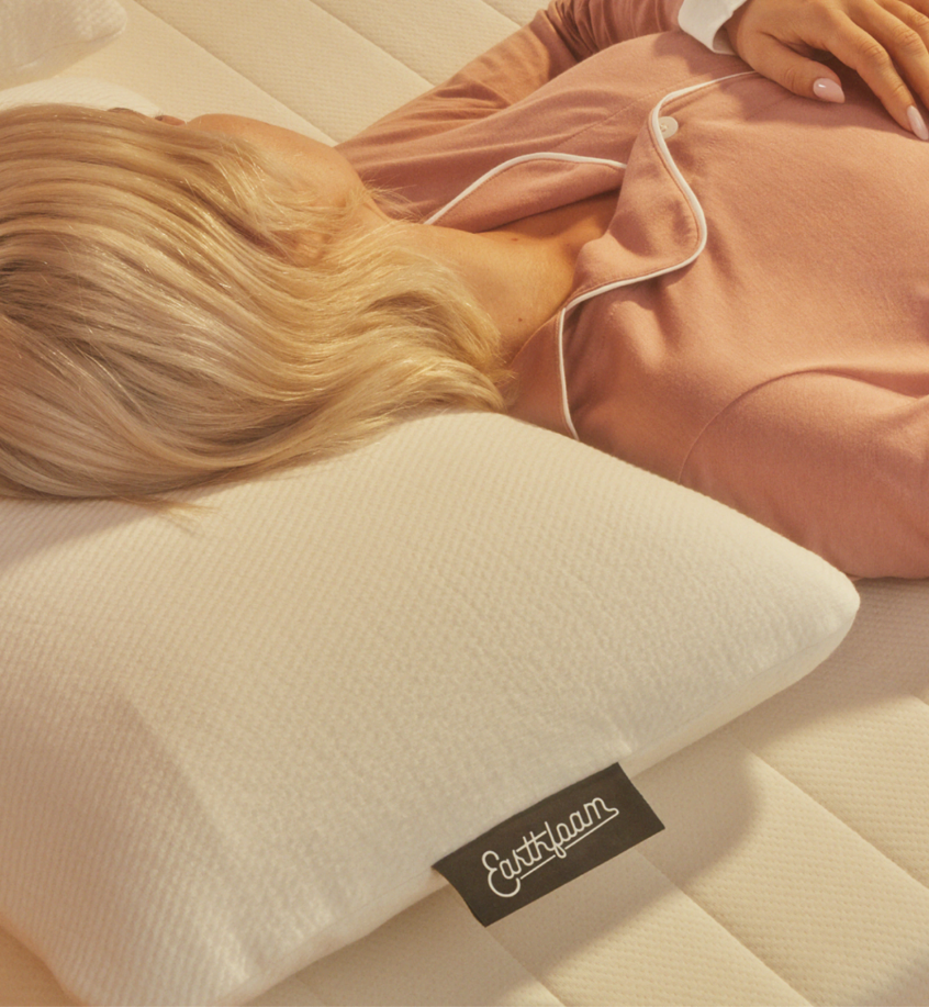 Person lying on their side in pink pajamas, resting on a white pillow with a visible "Earthfam" label, on a mattress.