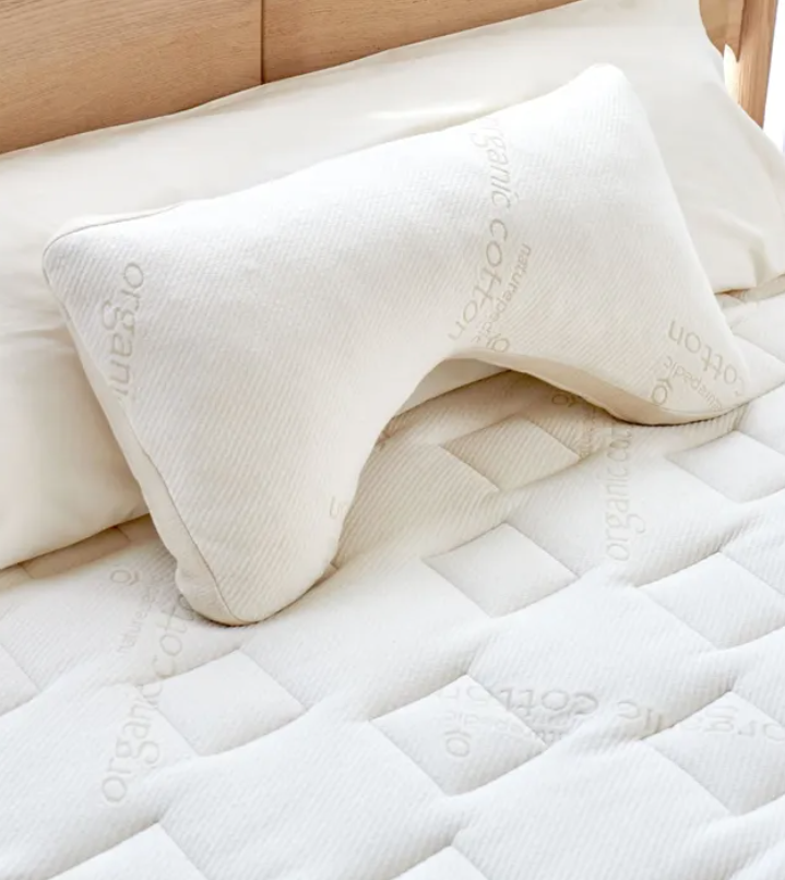 A white ergonomic pillow with "organic cotton" text lies on a matching quilted mattress and bedframe in a minimalist bedroom setting.
