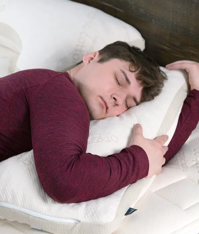 A person wearing a maroon long-sleeve shirt is sleeping facedown on a white pillow, hugging it with both arms.