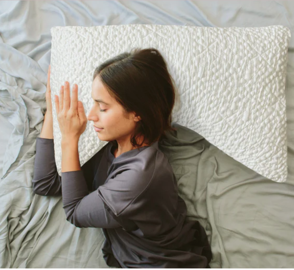 A woman lies on a bed with gray sheets, resting her head and hand on a large white textured pillow, eyes closed.