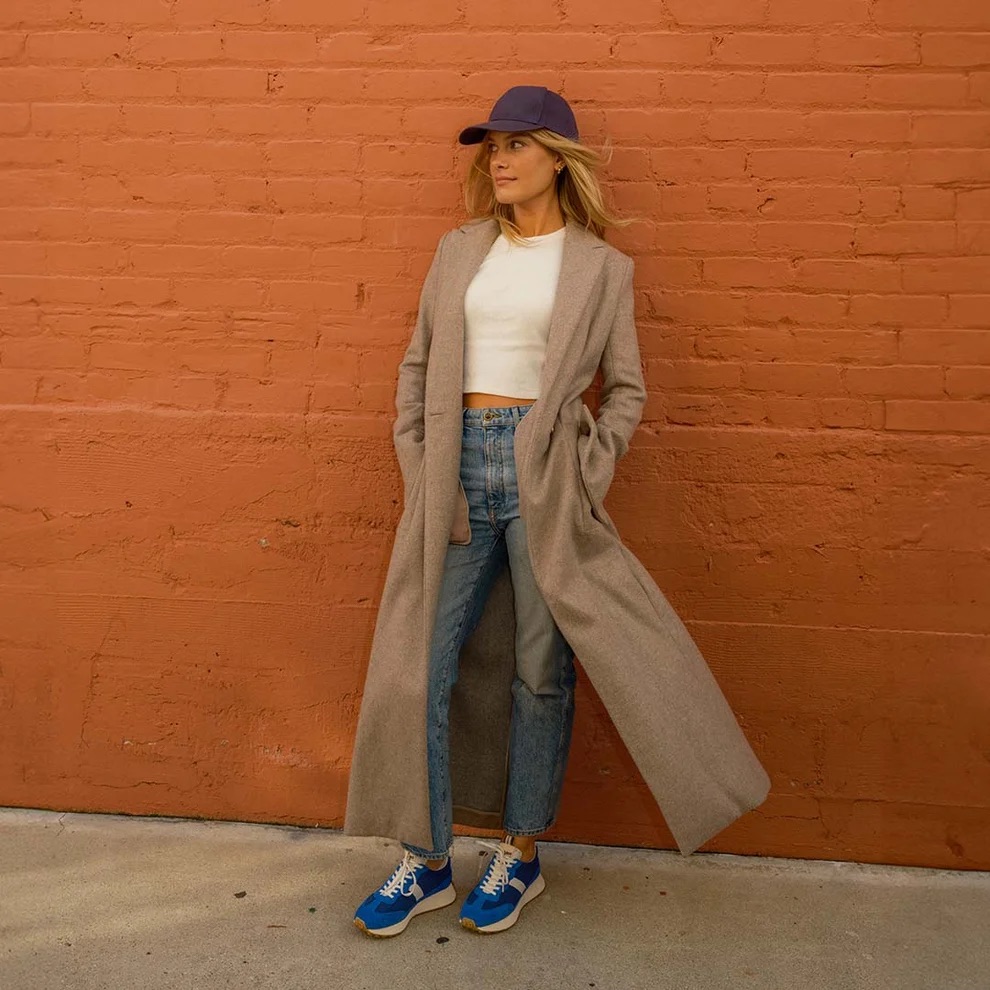 Woman in long beige coat, white crop top, blue jeans, blue sneakers, and a purple cap stands against an orange brick wall with hands in her pockets.