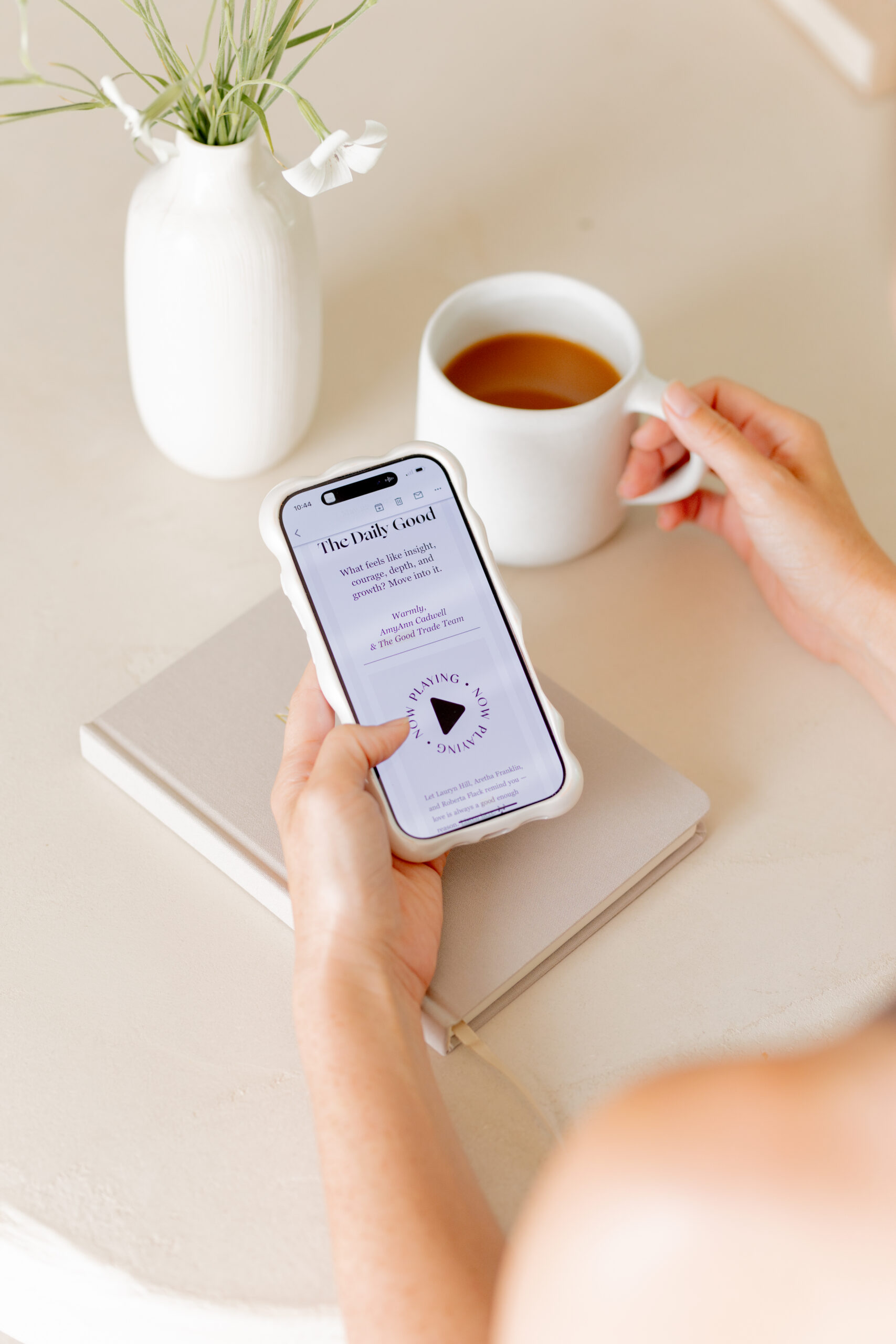 A person holding a cup of tea and reading a digital book on a smartphone, with a closed notebook and vase on a light-colored table.