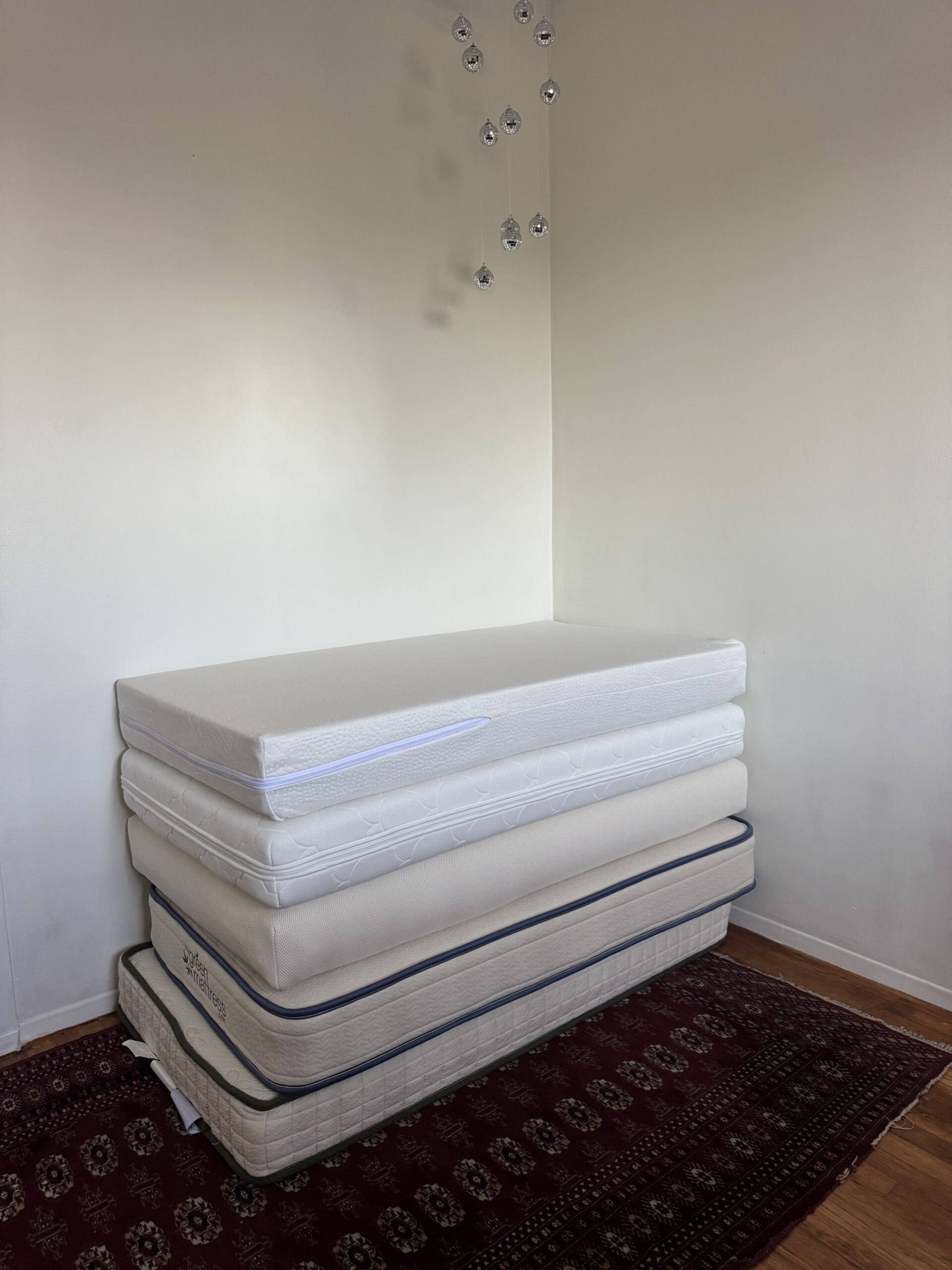 Four mattresses are stacked on top of each other in the corner of a room with a patterned rug, wooden floor, and a few small decorations on the wall.