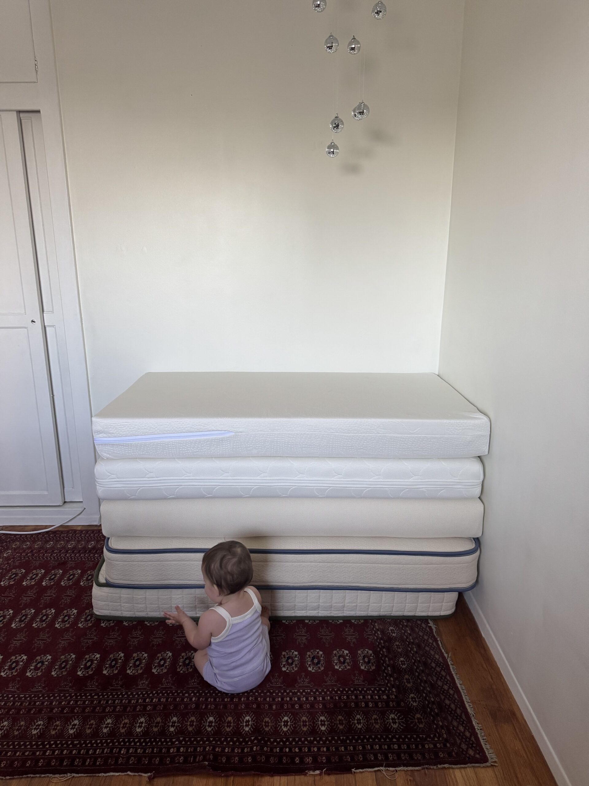 A baby sits on a red patterned rug in front of a stack of five mattresses against a white wall, with hanging crystal decorations above.