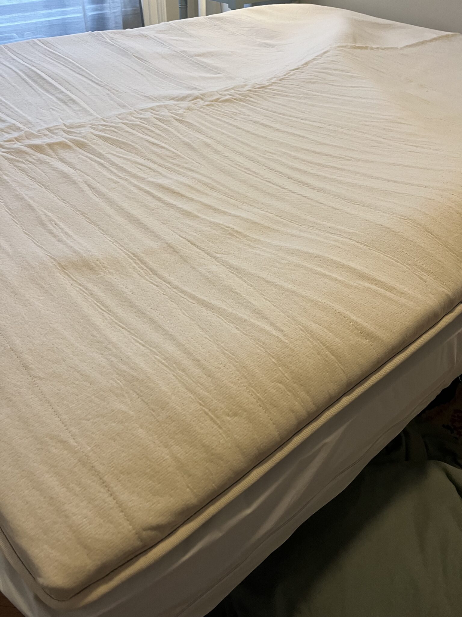 A close-up view of a bed with a beige fitted sheet, showing some wrinkles, in a well-lit room with window blinds in the background.