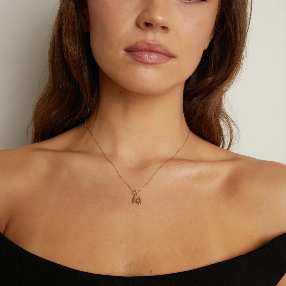 A woman with long brown hair wearing an off-shoulder black top and a thin gold necklace with a small letter "M" pendant.