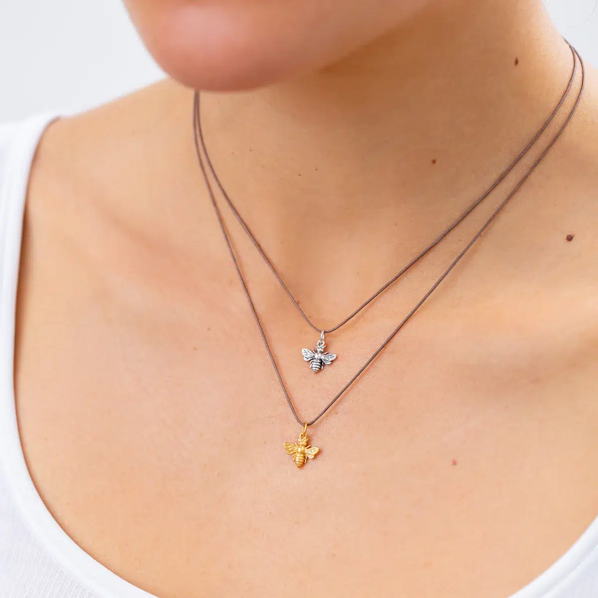 Close-up of a person wearing two thin necklaces, each with a small bee-shaped pendant, one gold and one silver, over a white tank top.