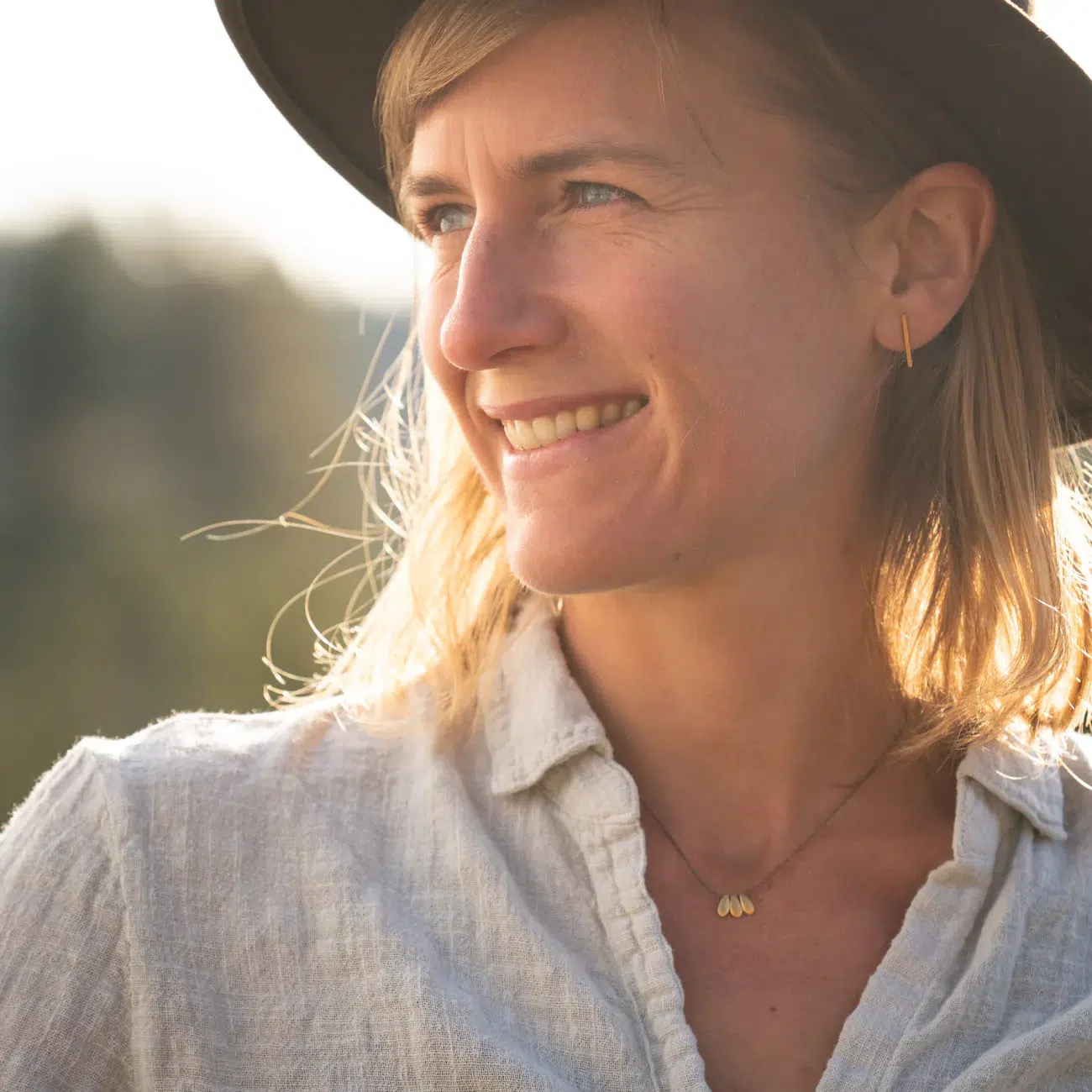 A woman wearing a hat and light-colored shirt smiles while looking to the side outdoors in soft natural light.