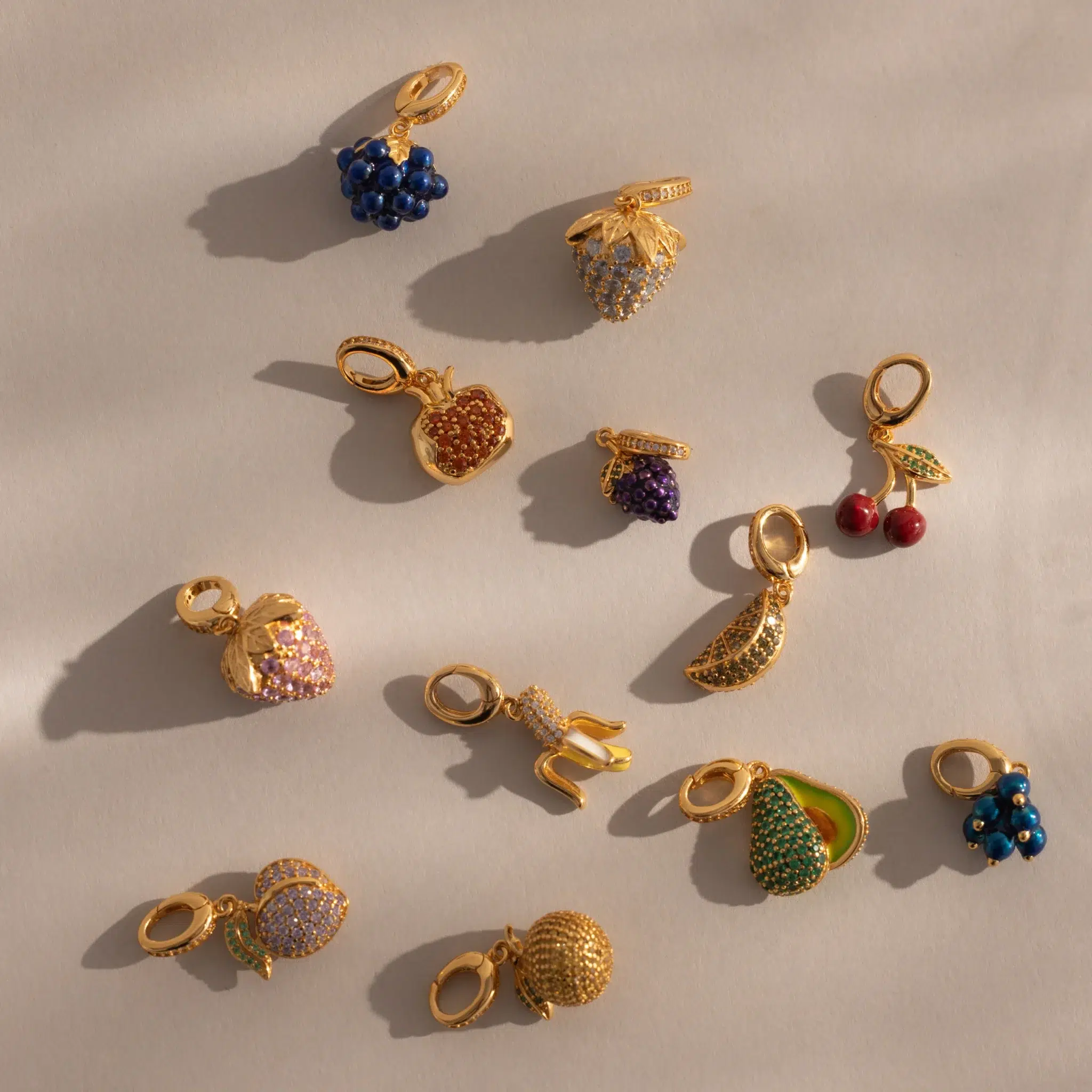 A collection of twelve small, gold charms shaped like fruits, an animal, and other objects, decorated with colorful gemstones, displayed on a neutral background.
