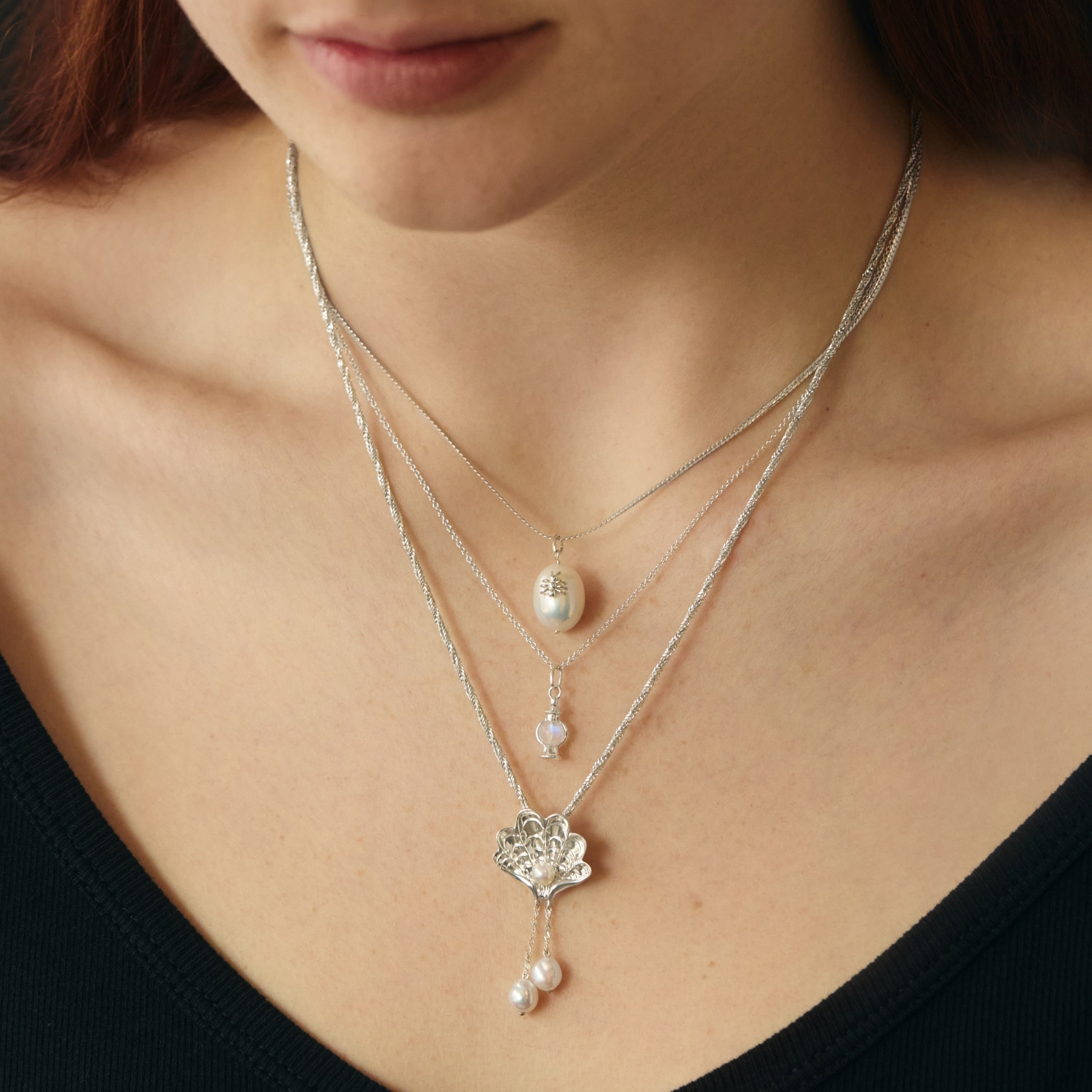 A woman wearing a black top displays three layered silver necklaces, each with a different pendant featuring pearls and a floral design.