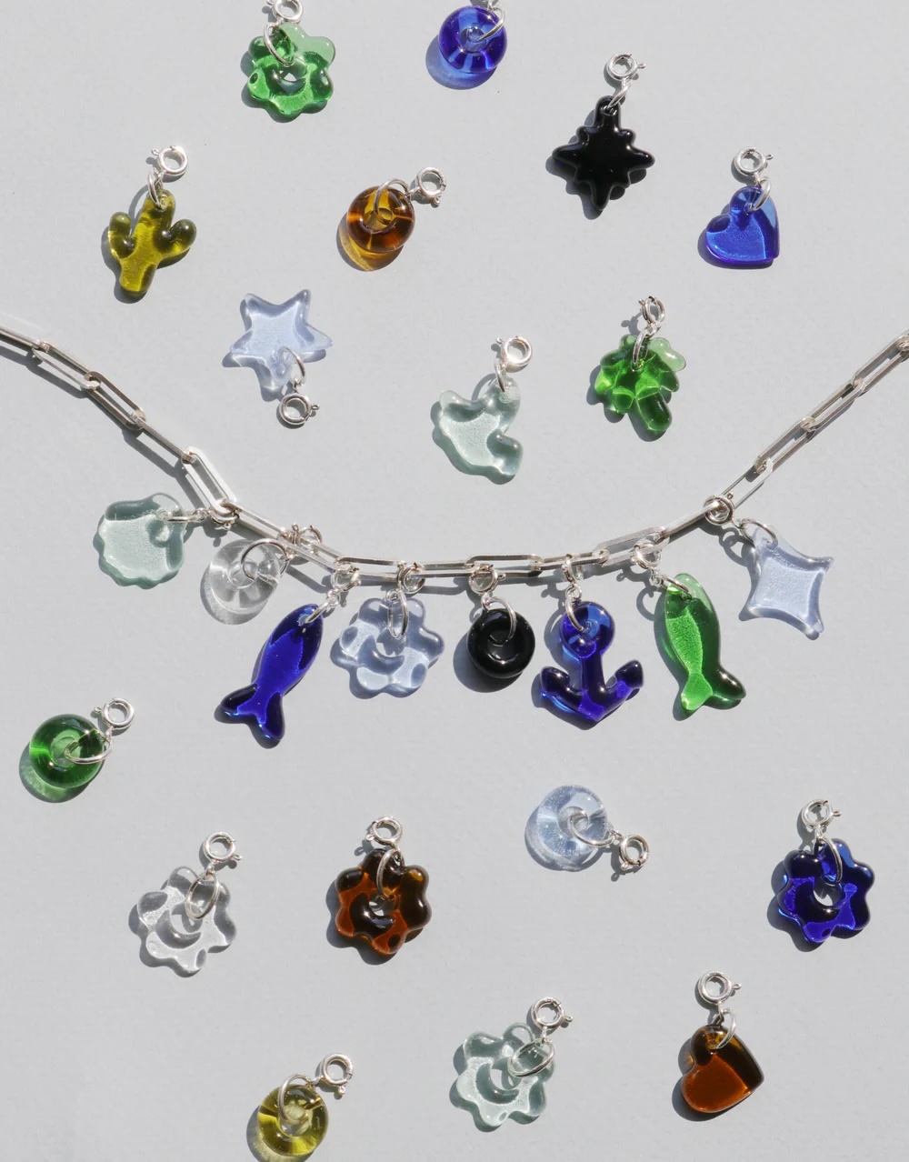 A silver chain with assorted colorful glass charms in various shapes, including animals and abstract forms, displayed on a light surface. Other loose charms are scattered around.