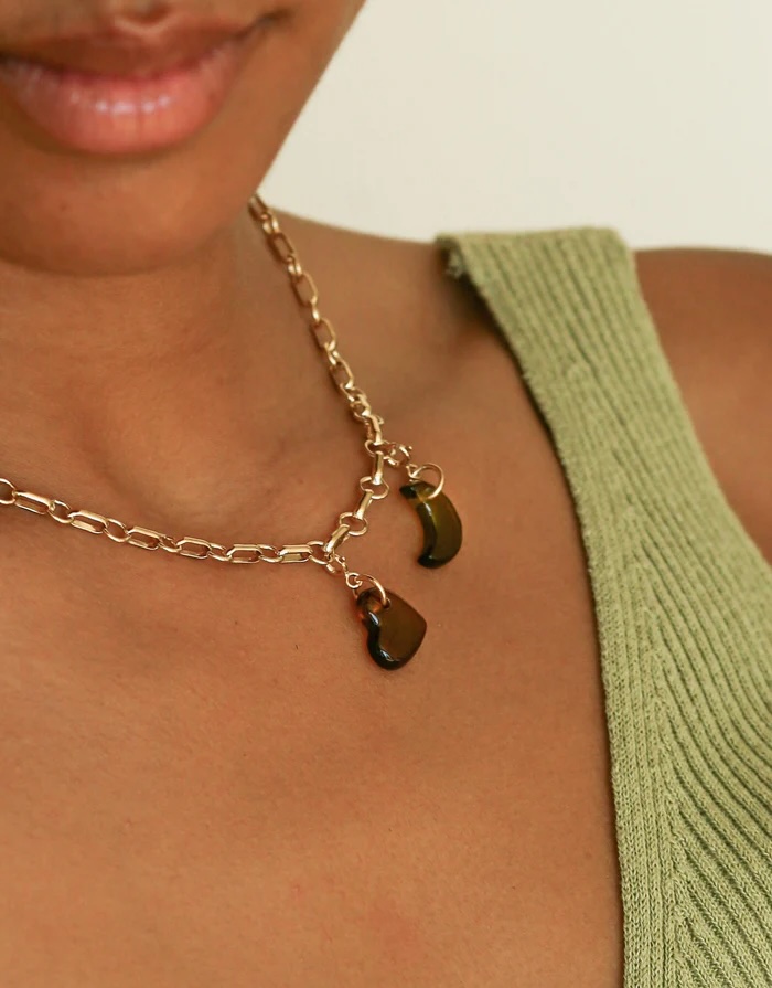 A person wearing a gold chain necklace with two dark, organic-shaped pendants, shown with a green textured sleeveless top.