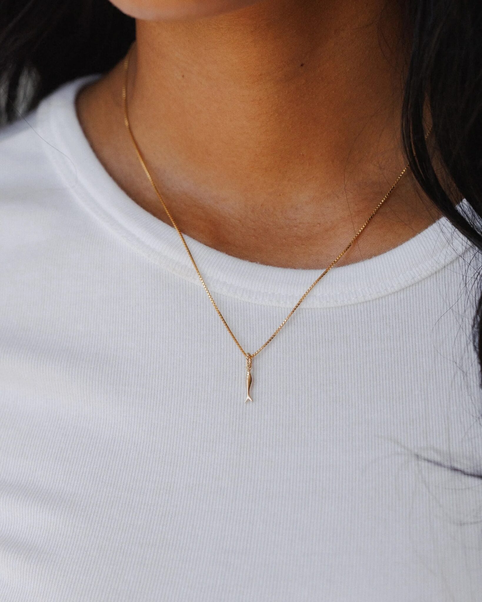 A person wearing a plain white ribbed shirt and a delicate gold chain necklace with a small, vertical pendant.