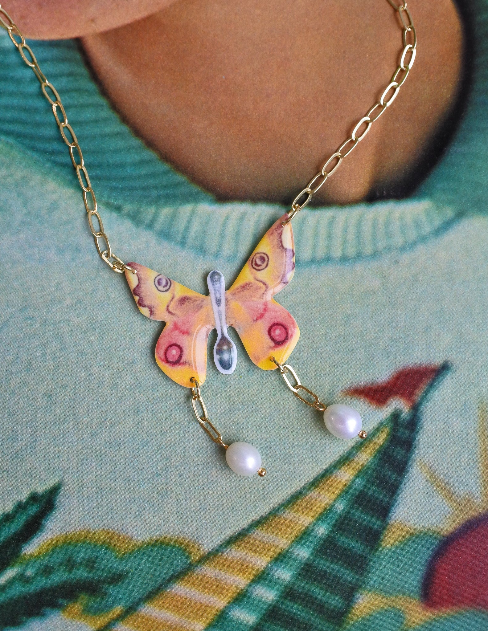 A gold chain necklace with a butterfly pendant and two dangling pearls, worn over a colorful patterned shirt.