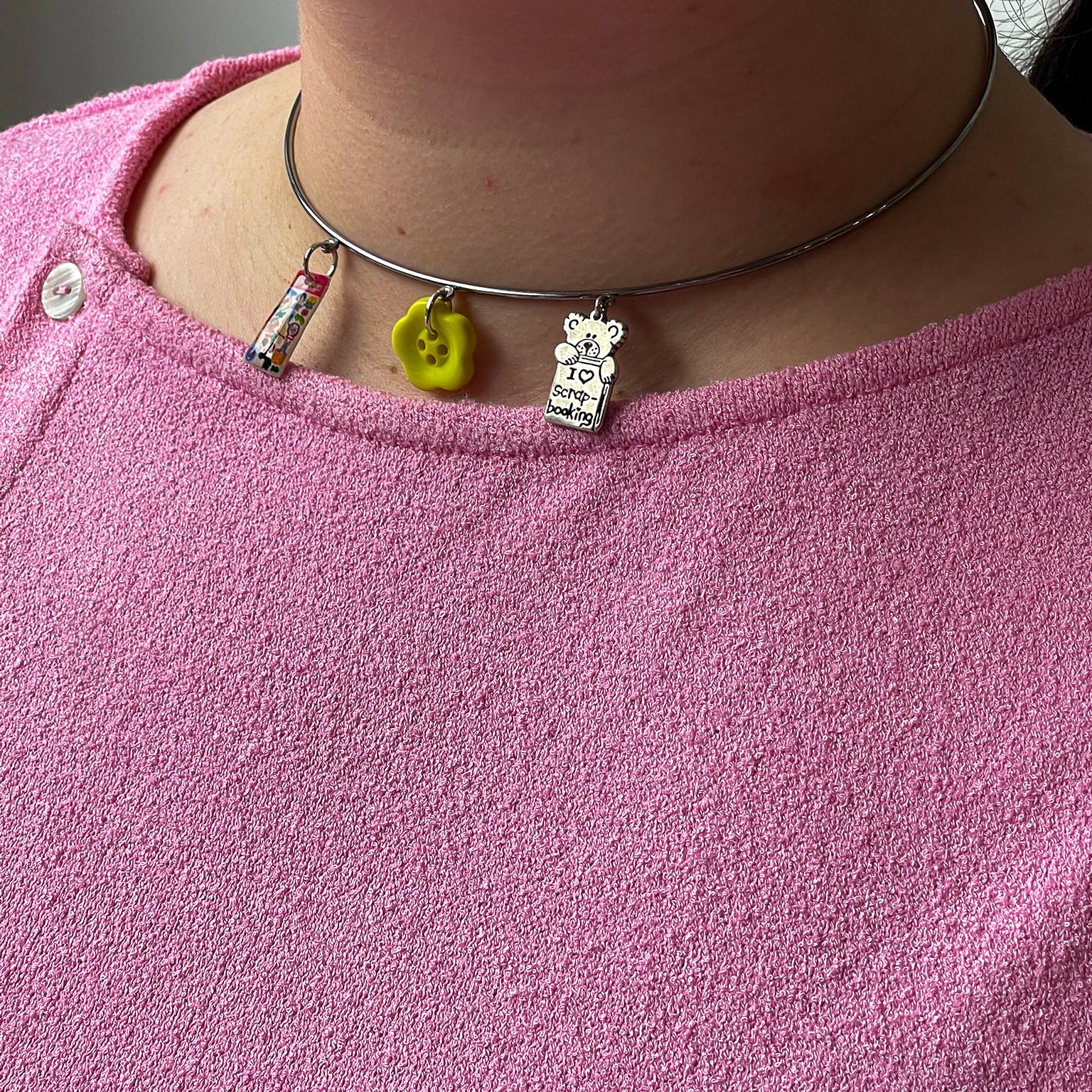A person wearing a pink textured shirt with a metal choker necklace featuring three charms: a mini book, a yellow smiley face, and a silver plaque that says "TO SERVE AND CORRECT.