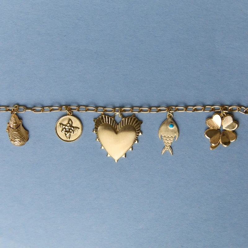 Gold charm bracelet with six charms: a hand, a round medallion, a heart, a fish, a fish tail with blue dot, and a four-leaf clover, on a light blue background.