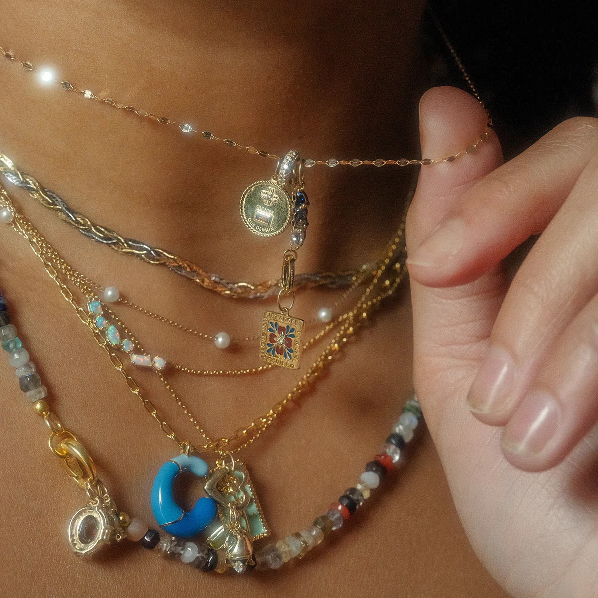 A close-up of a person wearing multiple layered necklaces with various colorful beads, charms, and pendants, and holding one chain between their fingers.