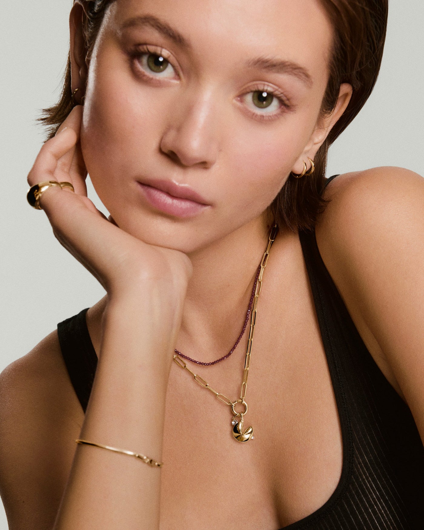 A person with short brown hair wears a black sleeveless top, gold jewelry including necklaces, a bracelet, and rings, and rests their face on their hand while looking at the camera.