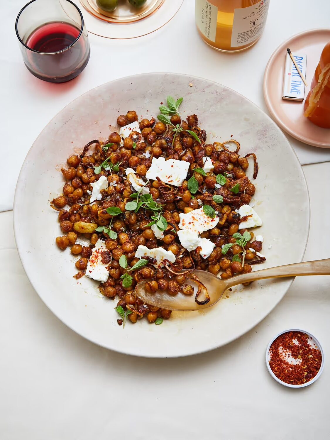 A large white bowl filled with roasted chickpeas, caramelized onions, crumbled cheese, and fresh herbs, with a serving spoon. A glass of red drink and condiments are nearby.