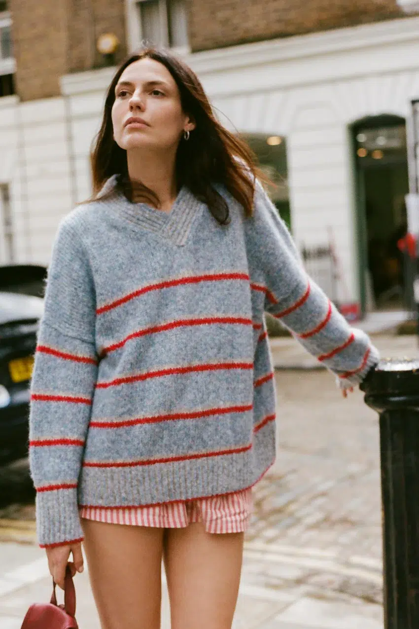 A woman stands outdoors wearing a gray sweater with red stripes and pink striped shorts, holding a red bag. The background features a street with buildings and parked cars.