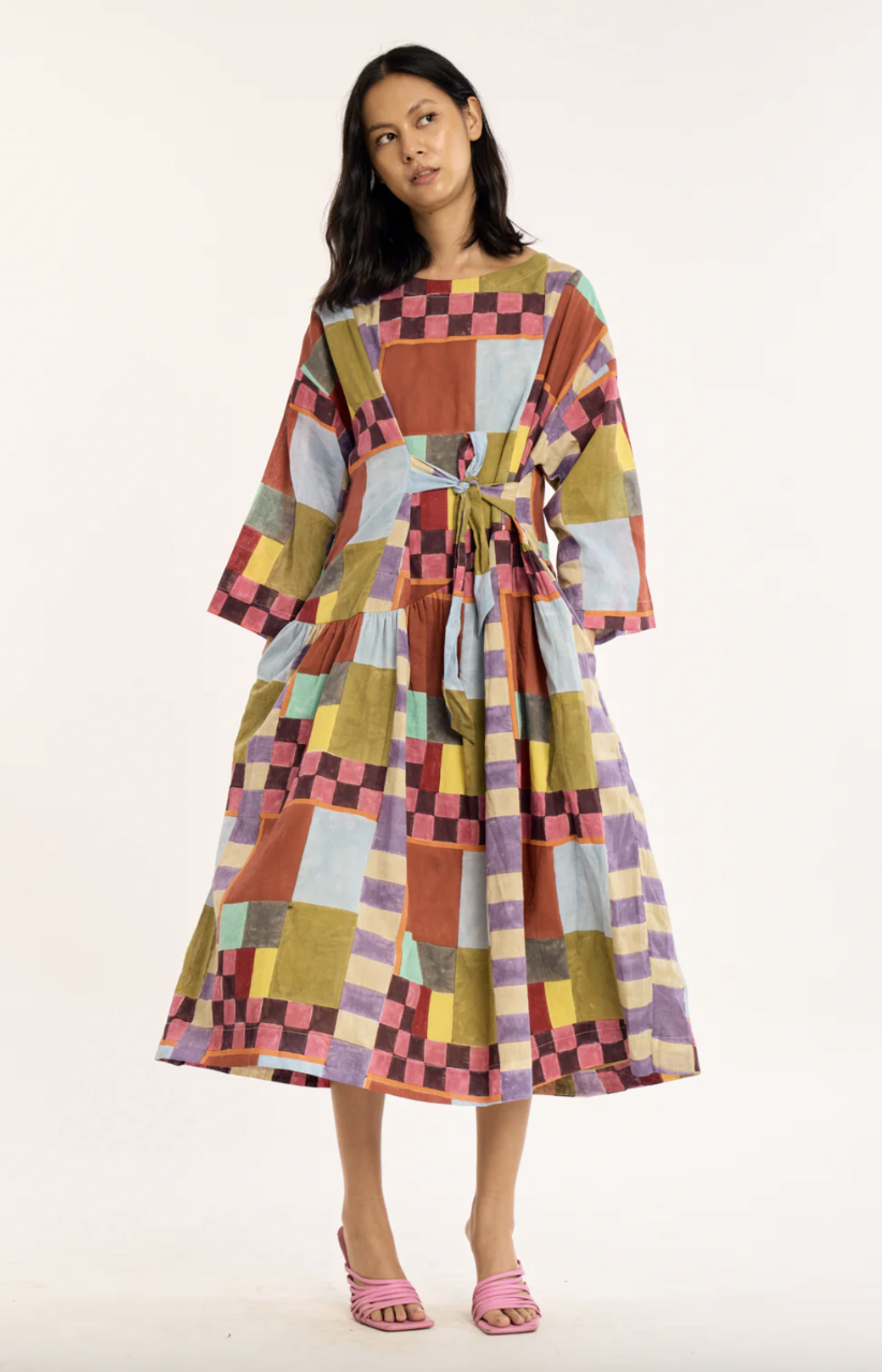 A woman stands wearing a loose, long-sleeved dress with a colorful patchwork pattern and pink heeled sandals against a plain background.