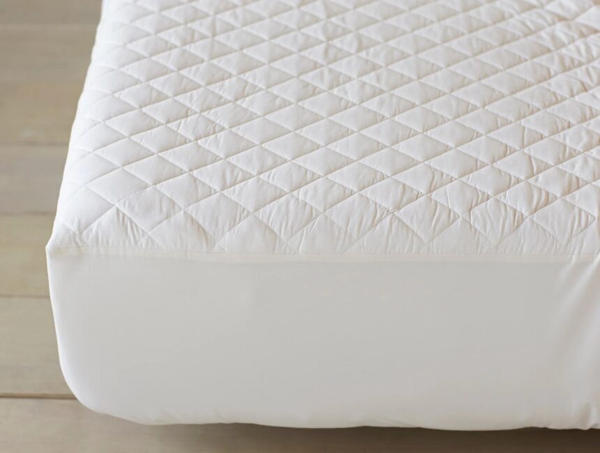 A close-up of a bed corner with a white, quilted mattress protector on a wooden floor.