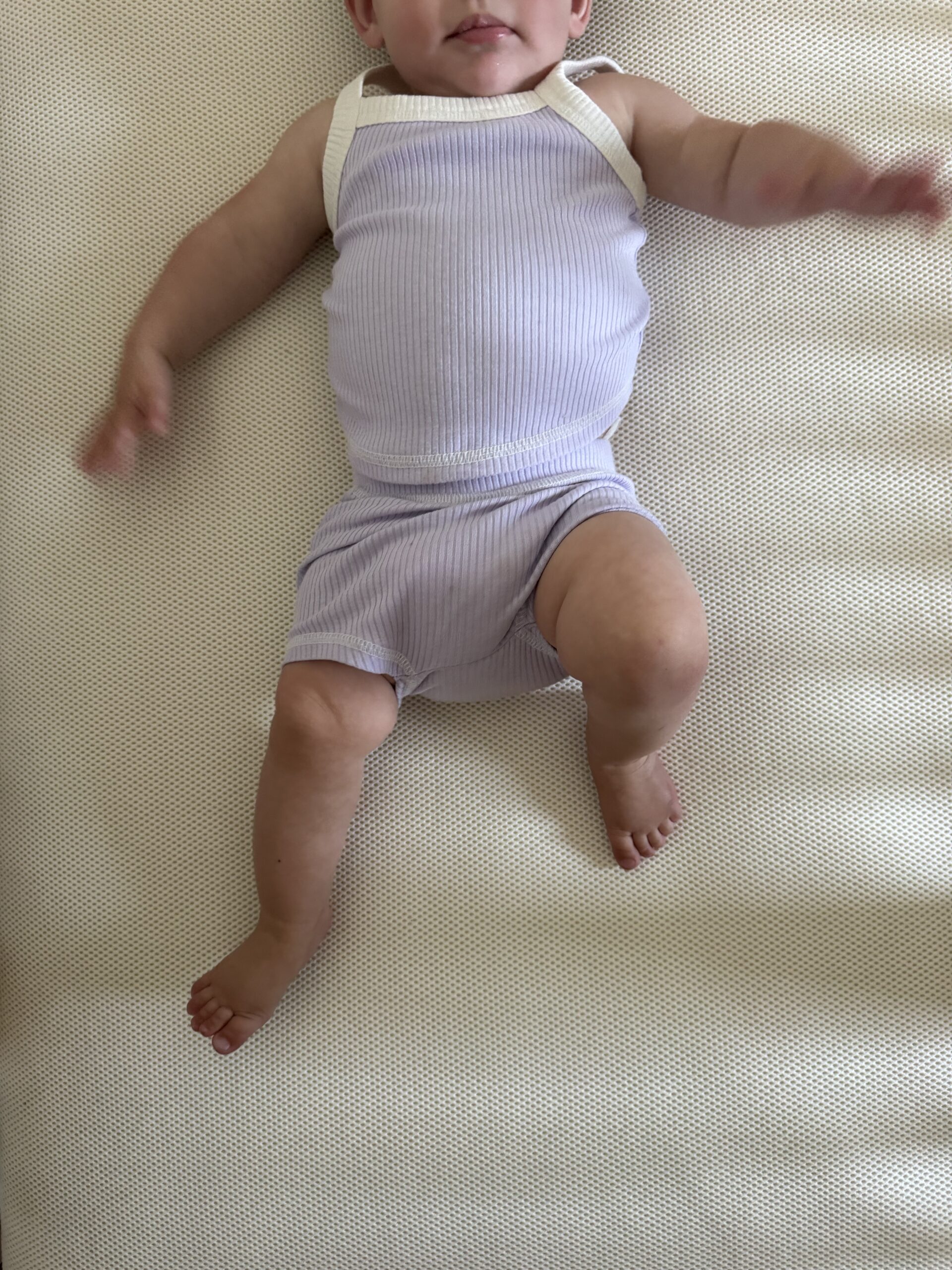 A baby wearing a light purple sleeveless outfit lies on a cream-colored textured surface, with arms and legs outstretched.