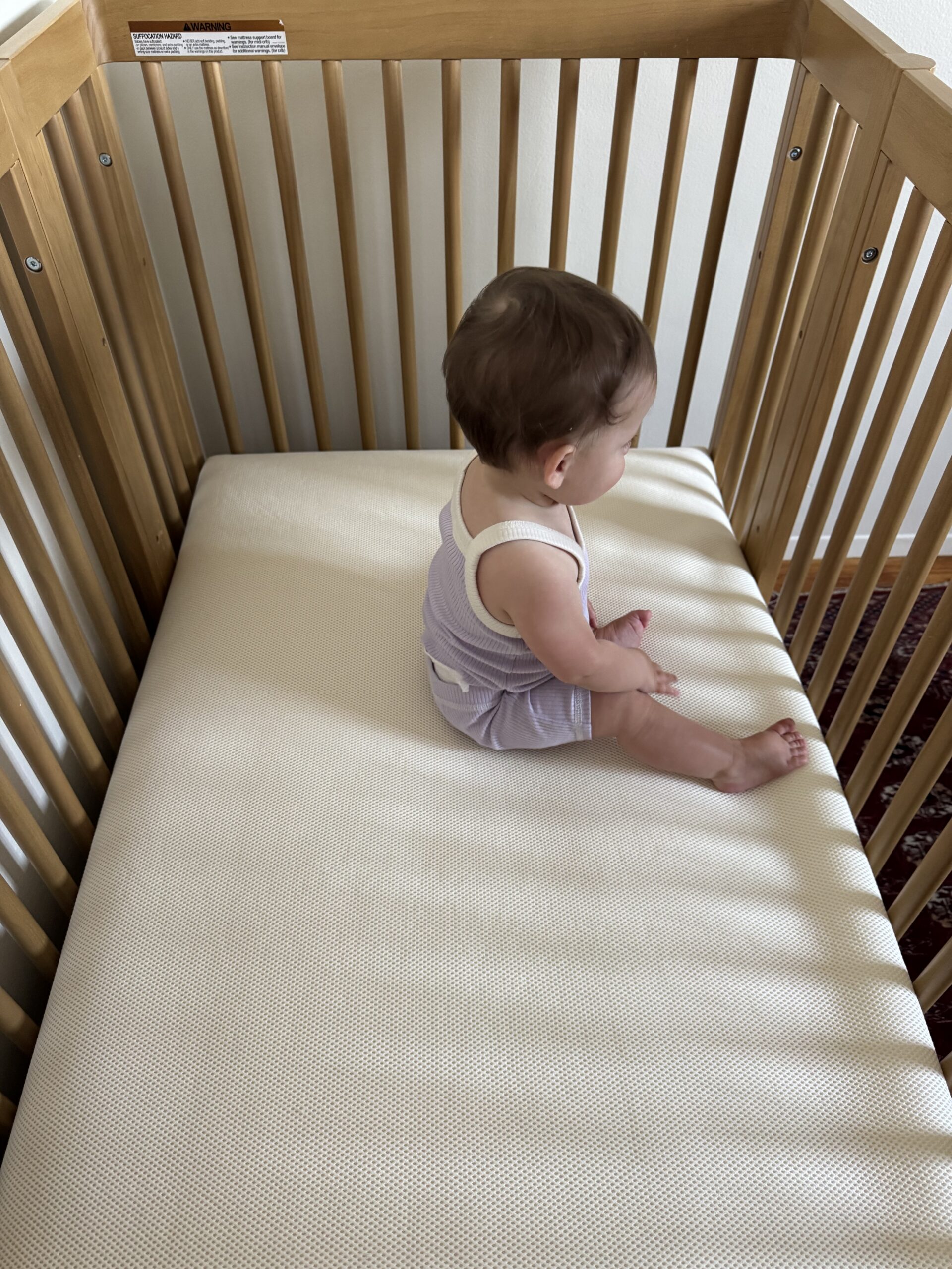 A baby in a light purple outfit sits upright in the corner of a wooden crib with a light-colored mattress.
