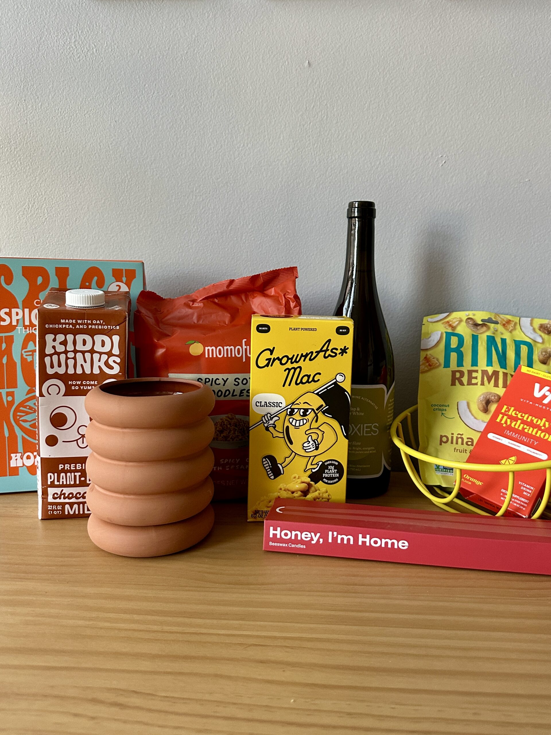 A variety of packaged food items, a wine bottle, a curved terracotta object, and a yellow ring are displayed on a wooden surface against a plain wall.