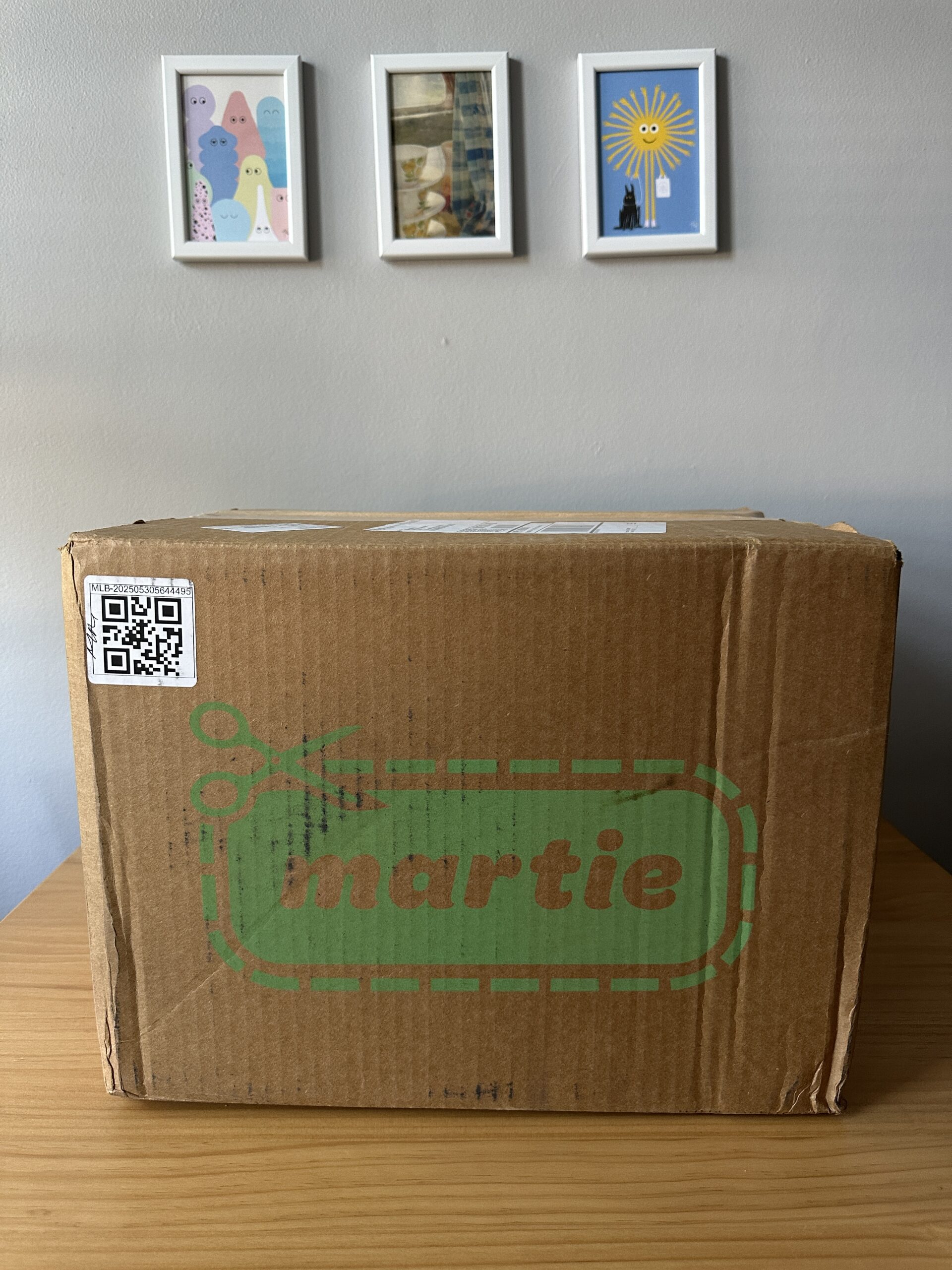 A sealed cardboard box with "martie" printed on it sits on a wooden table. Three framed pictures hang on the light gray wall behind it.