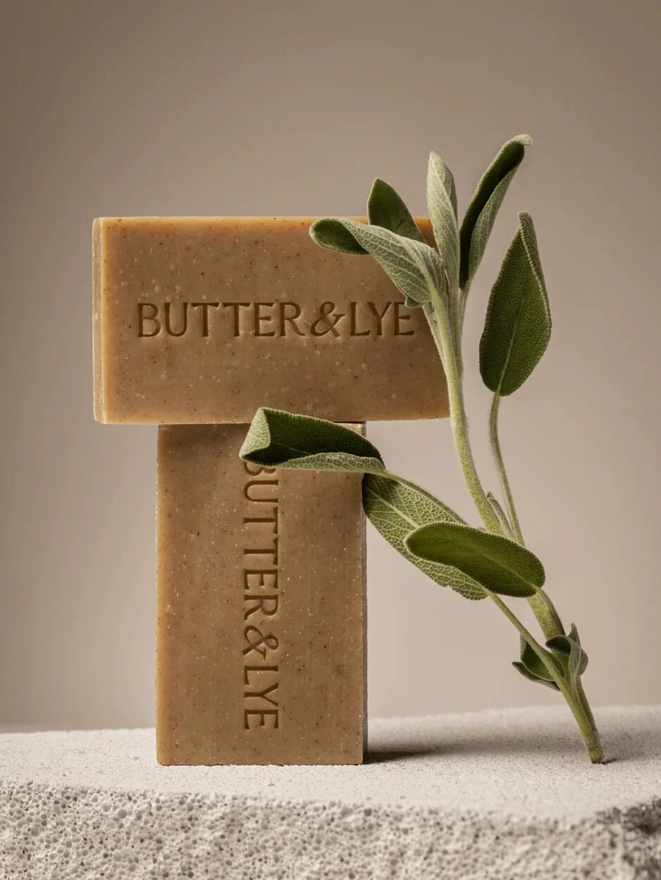 Two rectangular soap bars labeled "BUTTER&LYE" are stacked on a stone surface, with a sprig of fresh green sage leaning against them—perfect for elevating your self-care routine with just a few clicks through online shopping.