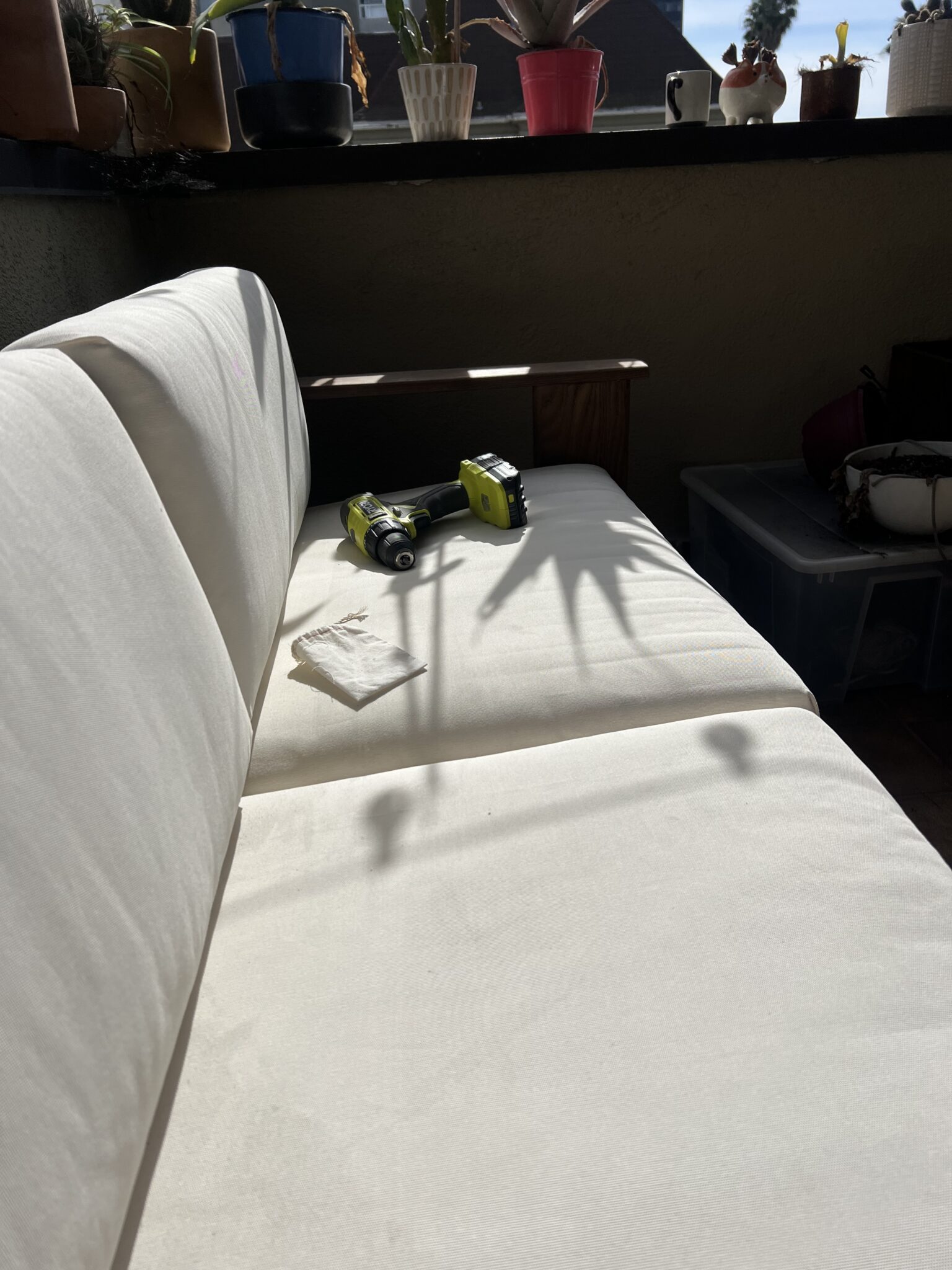 A white couch with a cordless drill, battery, and a paper towel on it; several potted plants line the windowsill in the background.