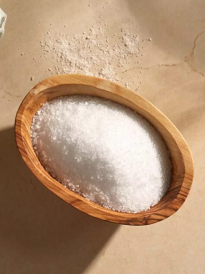 A wooden bowl filled with white bath salts sits on a beige surface next to a folded textured towel.