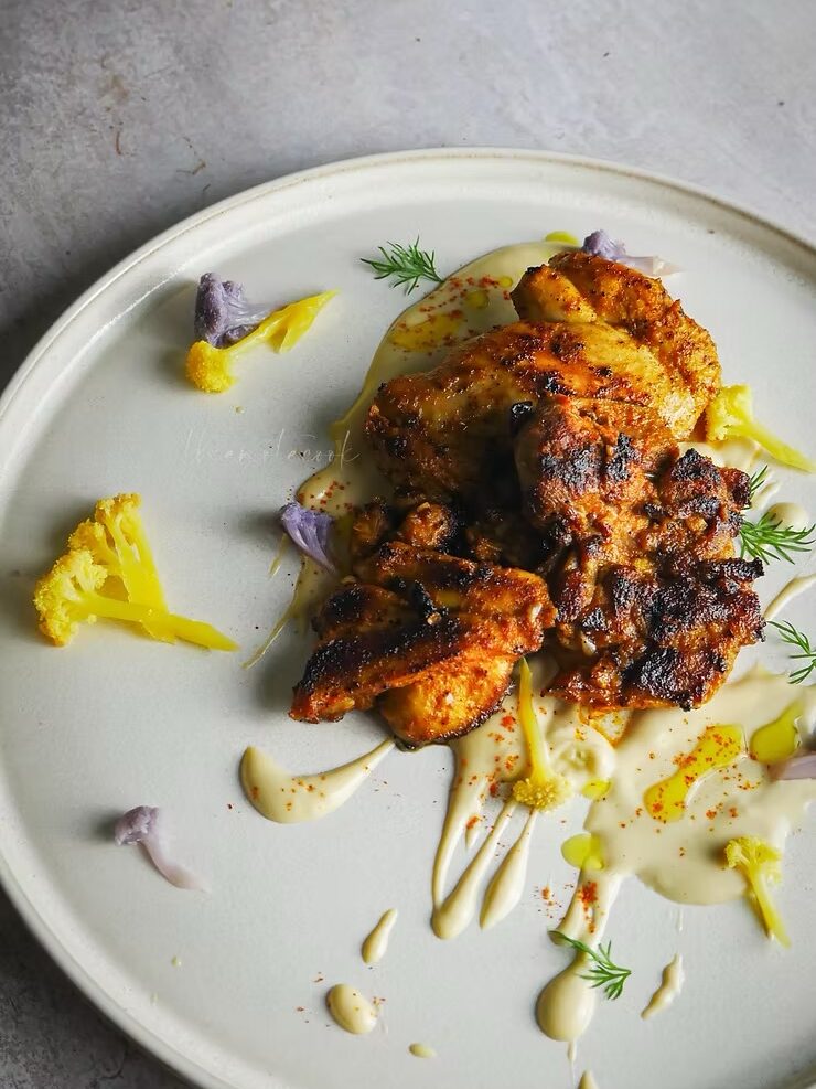 Grilled chicken pieces on a white plate with creamy sauce, garnished with small florets of yellow and purple cauliflower and fresh herbs.
